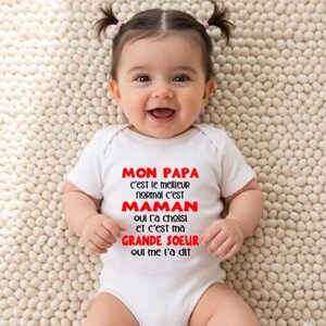 Peut inclure: Un bébé souriant portant un body blanc avec le texte "MON PAPA c'est le meilleur normal c'est MAMAN qui l'a choisi et c'est ma GRANDE SOEUR qui me l'a dit" en rouge et noir. Le bébé a deux petites couettes.