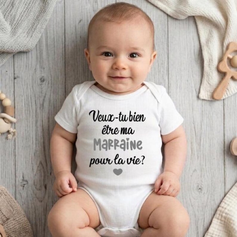 May include: A baby wearing a white onesie with the French text "Veux-tu bien &ecirc;tre ma Marraine pour la vie?" in black and gray lettering, with a small gray heart. The baby is lying on a light gray wooden surface.