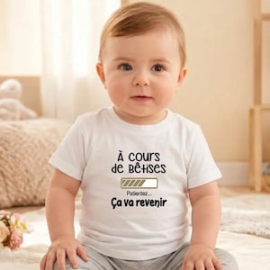 Peut inclure: Un bébé porte un t-shirt blanc avec du texte noir. Le texte dit "À cours de bêtises" au-dessus d'une barre de chargement, suivi de "Patientez... Ça va revenir". Le bébé est assis et regarde la caméra.