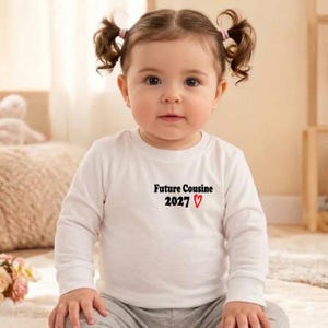 Peut inclure: Un bébé portant un t-shirt blanc à manches longues avec le texte "Future Cousin 2027" et un cœur rouge. Le bébé a les cheveux bruns en couettes et regarde la caméra.