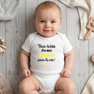 May include: A smiling baby wearing a white onesie with the French text "Veux-tu bien &ecirc;tre mon parrain pour la vie?" in black and yellow. The baby is lying on a light gray wooden surface, surrounded by baby toys.
