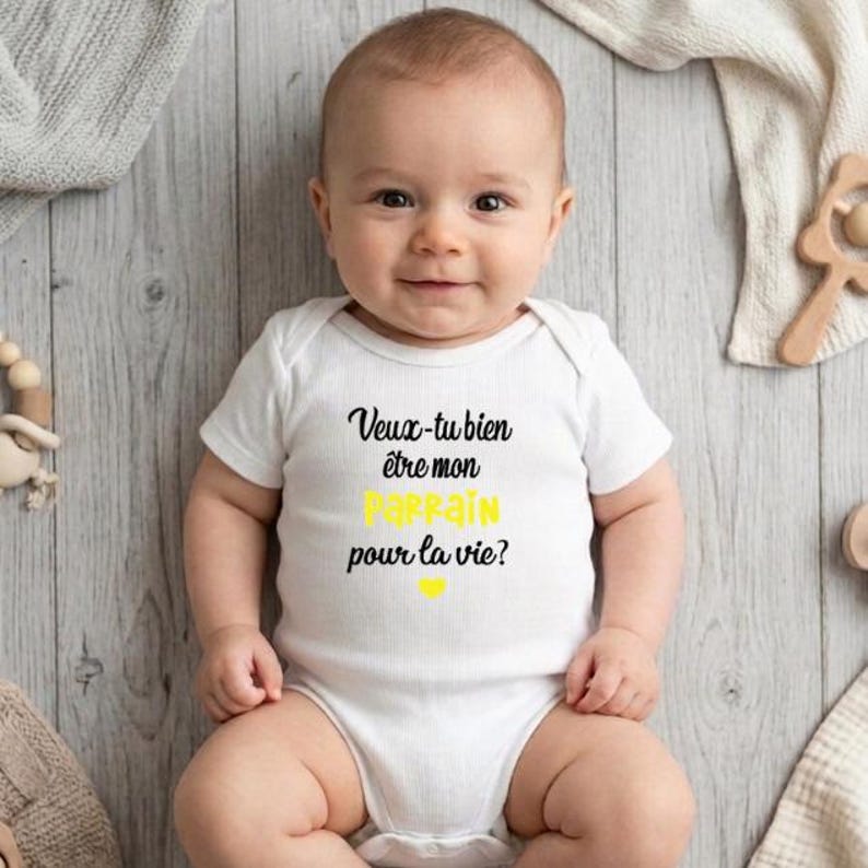 May include: A smiling baby wearing a white onesie with the French text "Veux-tu bien &ecirc;tre mon parrain pour la vie?" in black and yellow. The baby is lying on a light gray wooden surface, surrounded by baby toys.