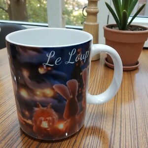 Peut inclure: Mug en céramique blanche avec une illustration enveloppante représentant une scène nocturne avec des personnages de dessins animés et le texte "Le Loup" en écriture manuscrite. La tasse a une anse blanche et un intérieur bleu clair.
