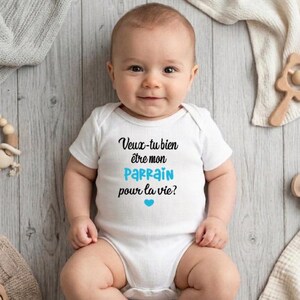 May include: A smiling baby wearing a white onesie with the French text "Veux-tu bien &ecirc;tre mon parrain pour la vie?" in black and blue. A blue heart is below the text. The baby is lying on a light wood-grain surface.