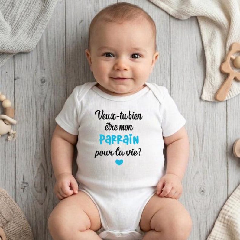 May include: A smiling baby wearing a white onesie with the French text "Veux-tu bien &ecirc;tre mon parrain pour la vie?" in black and blue. A blue heart is below the text. The baby is lying on a light wood-grain surface.