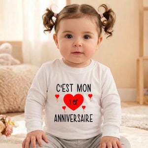 May include: A baby wearing a white long-sleeved shirt with the text "C'EST MON 1er ANNIVERSAIRE" in black and red. The shirt features a large red heart and smaller red hearts. The baby has brown hair styled in pigtails.