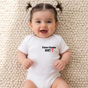 Peut inclure: Un bébé souriant portant un body blanc avec le texte "Future Cousine 2027" et un cœur rouge. Le bébé est allongé sur une surface texturée blanc cassé.