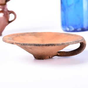 May include: A small, terracotta bowl with a handle. The bowl is slightly angled and has a rough, textured surface.
