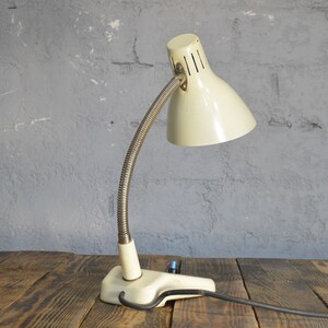 K&ouml;nnte beinhalten: Eine wei&szlig;e Vintage-Tischlampe mit flexiblem Arm und einer Klemmbasis. Die Lampe hat einen wei&szlig;en Metallschirm mit einer silbernen Metallbasis.