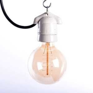 May include: A vintage-style light bulb with a warm, amber glow. The bulb is round and suspended from a white ceramic fixture and a black wire. The filament is visible inside the glass. The light fixture is hanging from a metal hook.