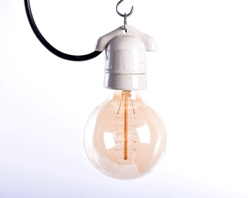 May include: A vintage-style light bulb with a warm, amber glow. The bulb is round and suspended from a white ceramic fixture and a black wire. The filament is visible inside the glass. The light fixture is hanging from a metal hook.