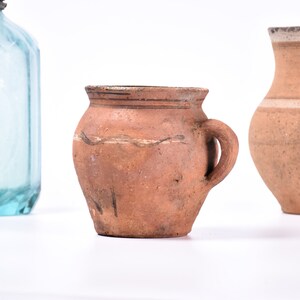 Pode incluir: Três vasos de cerâmica, dois são marrons com uma textura áspera e um é um vaso marrom menor com uma alça. O vaso marrom maior tem uma borda branca.