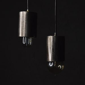 Könnte beinhalten: Zwei zylindrische Pendelleuchten aus dunklem Metall hängen vor einem schwarzen Hintergrund. Die Leuchten haben eine strukturierte Oberfläche und freiliegende Glühbirnen. Eine Glühbirne hat einen U-förmigen Glühfaden, die andere eine runde Glühbirne.