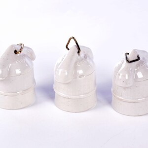 May include: Three off-white ceramic electrical insulators with a rounded top and a metal loop for hanging. The insulators have a cylindrical base and are arranged against a white background.
