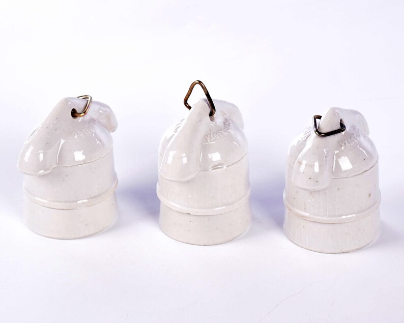 May include: Three off-white ceramic electrical insulators with a rounded top and a metal loop for hanging. The insulators have a cylindrical base and are arranged against a white background.