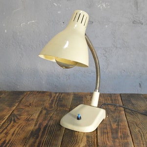 K&ouml;nnte beinhalten: Eine wei&szlig;e Vintage-Tischlampe mit flexiblem Arm und wei&szlig;em Schirm. Die Lampe hat einen runden Sockel mit einem blauen Schalter.