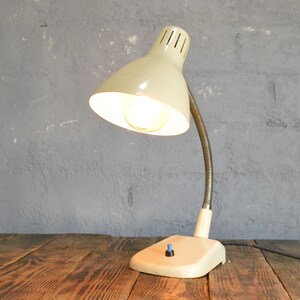 K&ouml;nnte beinhalten: Eine wei&szlig;e Vintage-Tischlampe mit flexiblem Arm und wei&szlig;em Schirm. Die Lampe hat eine eingeschaltete Gl&uuml;hbirne und steht auf einem Holztisch.