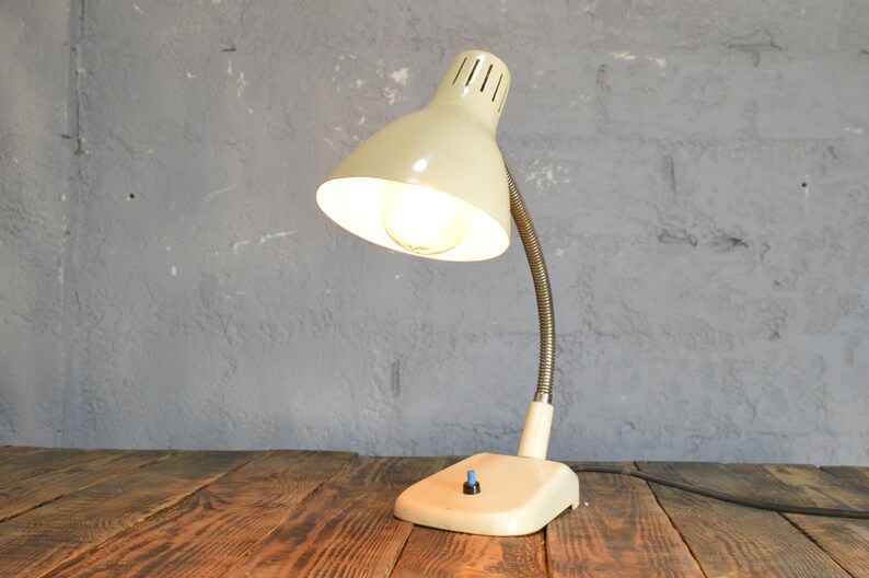 K&ouml;nnte beinhalten: Eine wei&szlig;e Vintage-Tischlampe mit flexiblem Arm und wei&szlig;em Schirm. Die Lampe hat eine eingeschaltete Gl&uuml;hbirne und steht auf einem Holztisch.