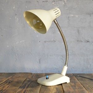 K&ouml;nnte beinhalten: Eine wei&szlig;e Vintage-Tischlampe mit flexiblem Arm und einem wei&szlig;en Ein-/Ausschalter. Die Lampe hat einen wei&szlig;en Schirm mit einem Metallring oben.