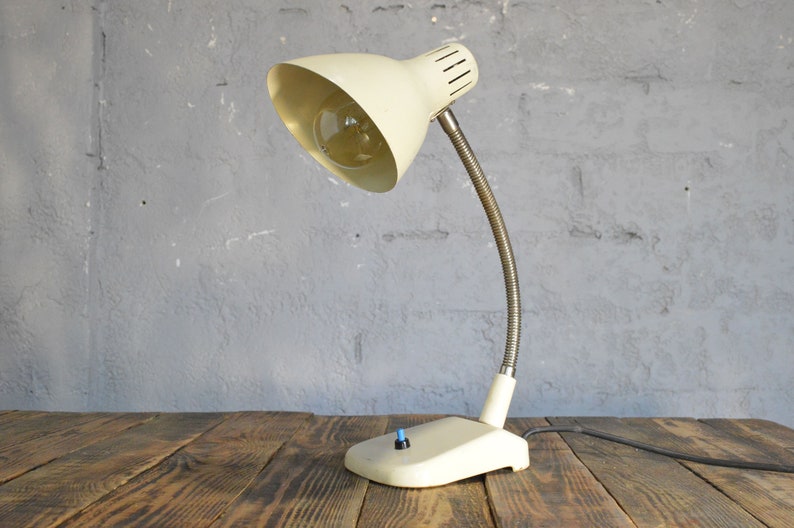 K&ouml;nnte beinhalten: Eine wei&szlig;e Vintage-Tischlampe mit flexiblem Arm und einem wei&szlig;en Ein-/Ausschalter. Die Lampe hat einen wei&szlig;en Schirm mit einem Metallring oben.