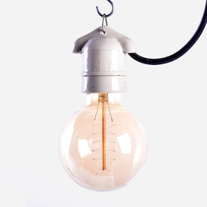 May include: A spherical, vintage-style light bulb with a warm amber glow hangs from a white ceramic socket and a black cord. The bulb's filament is visible, and the design evokes a classic, industrial aesthetic. The light fixture is suspended against a white background.