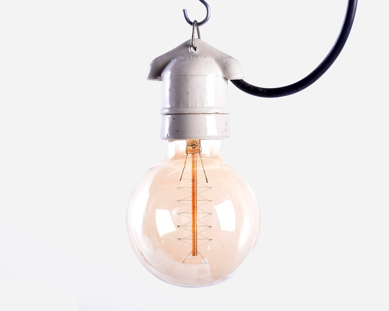 May include: A vintage-style light bulb with a warm, amber glow hangs from a white ceramic socket and a black cord. The bulb's filament is visible, and the design evokes a classic, industrial aesthetic. The light fixture is suspended against a white background.