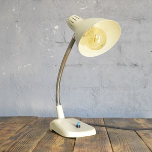 K&ouml;nnte beinhalten: Eine wei&szlig;e Vintage-Tischlampe mit einem flexiblen Schwanenhalsarm und einem wei&szlig;en Schirm. Die Lampe hat eine Gl&uuml;hbirne und ein schwarzes Kabel.