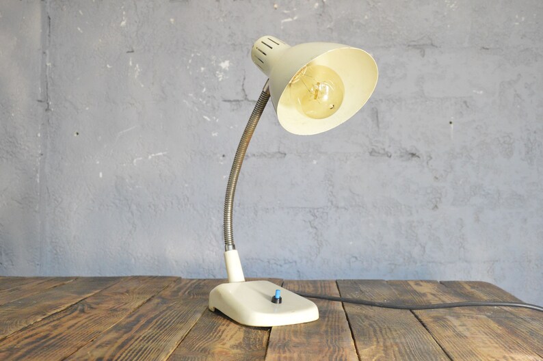 K&ouml;nnte beinhalten: Eine wei&szlig;e Vintage-Tischlampe mit einem flexiblen Schwanenhalsarm und einem wei&szlig;en Schirm. Die Lampe hat eine Gl&uuml;hbirne und ein schwarzes Kabel.