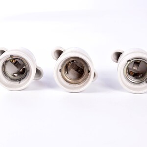 May include: Three white ceramic light bulb sockets with metal contacts and small loop handles. The sockets are arranged in a row against a white background, showcasing their interior details and design.