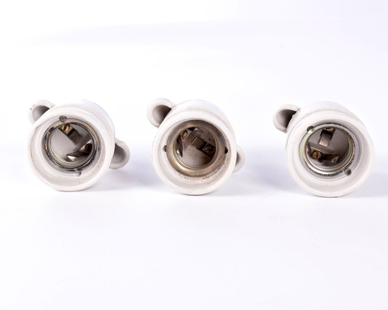 May include: Three white ceramic light bulb sockets with metal contacts and small loop handles. The sockets are arranged in a row against a white background, showcasing their interior details and design.