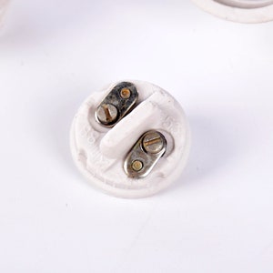 May include: A close-up of a white ceramic electrical component with two metal contact points secured by screws. The component has a rounded shape with a raised central section and is set against a white background.