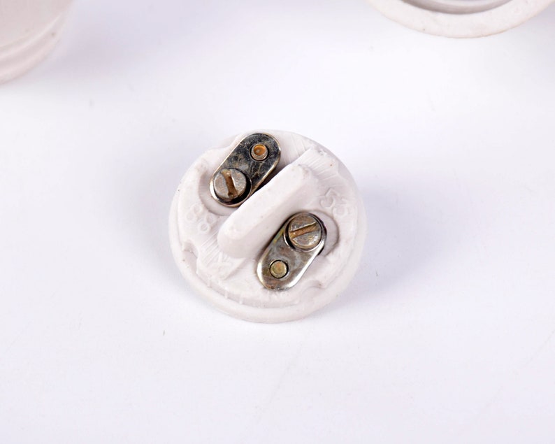 May include: A close-up of a white ceramic electrical component with two metal contact points secured by screws. The component has a rounded shape with a raised central section and is set against a white background.