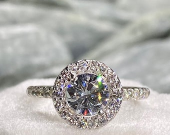 Könnte beinhalten: Ein silberner Verlobungsring mit einem runden, klaren Mittelstück, das von einem Heiligenschein aus kleineren Diamanten umgeben ist. Der Ring liegt in einer weißen Samt-Ringbox.