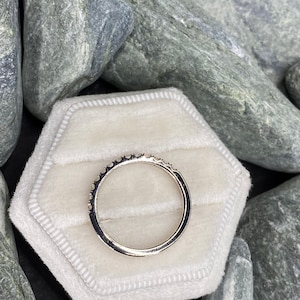 May include: A silver band ring with small diamonds set in a pave setting. The ring is displayed on a white velvet ring box.
