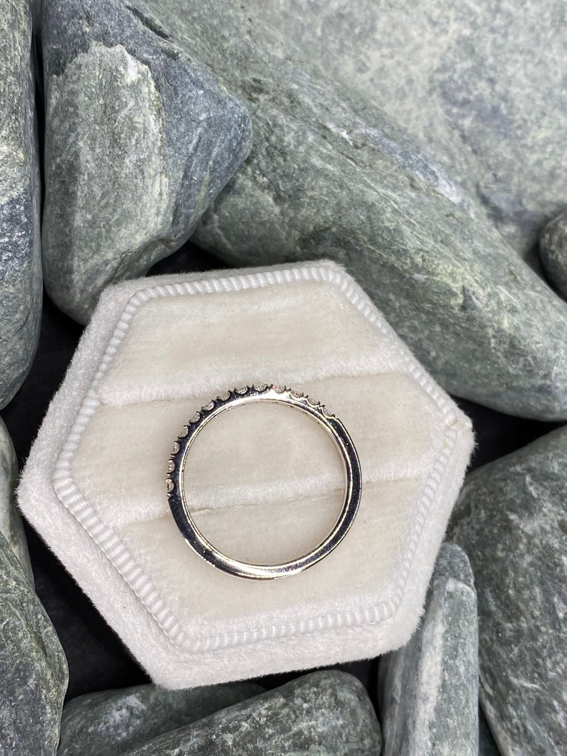 May include: A silver band ring with small diamonds set in a pave setting. The ring is displayed on a white velvet ring box.