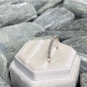 May include: A silver band ring with a row of small, clear gemstones set in a pave setting. The ring is displayed in a white velvet ring box.