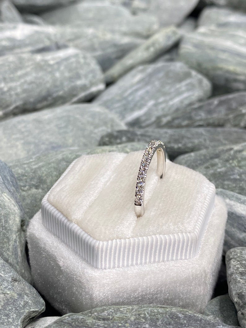 May include: A silver band ring with a row of small, clear gemstones set in a pave setting. The ring is displayed in a white velvet ring box.