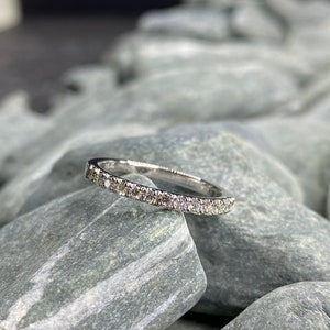 May include: A thin silver band with a row of small diamonds set in a pave setting. The ring is resting on a pile of gray stones.