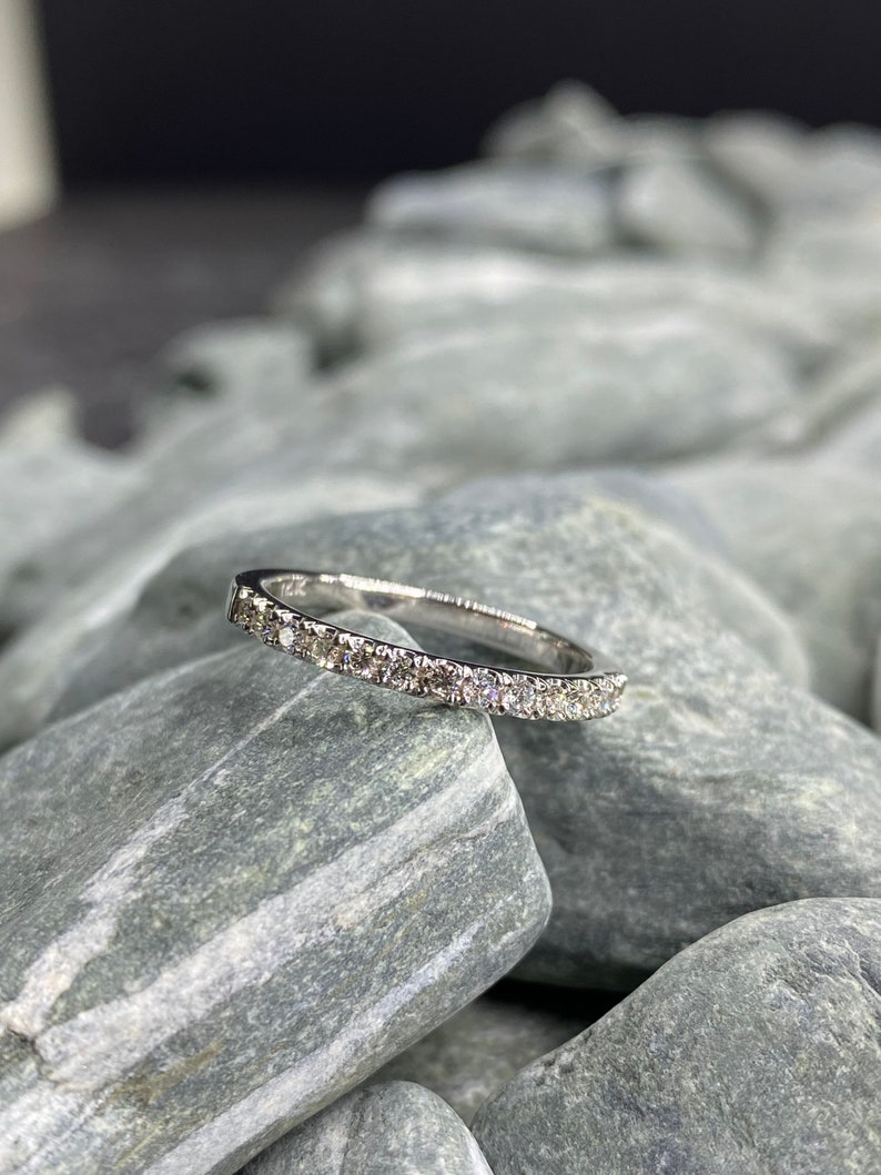 May include: A thin silver band with a row of small diamonds set in a pave setting. The ring is resting on a pile of gray stones.
