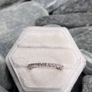May include: A thin silver band with a row of small, clear diamonds set in a pave setting. The ring is displayed in a white velvet ring box.
