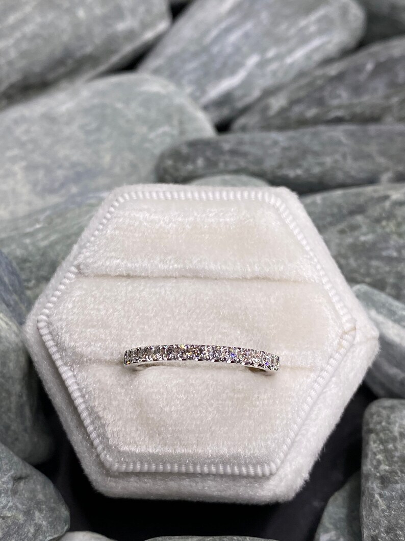 May include: A thin silver band with a row of small, clear diamonds set in a pave setting. The ring is displayed in a white velvet ring box.