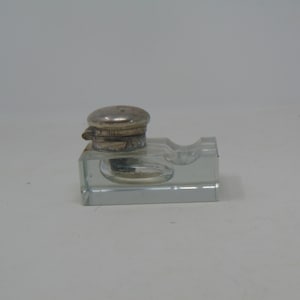 Peut inclure: Un petit encrier en verre transparent avec un couvercle en argent. L'encrier a une base rectangulaire et un dessus rond. Le couvercle a une ouverture à charnière.