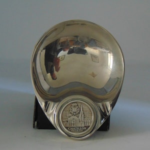 May include: A silver-colored, round, shallow dish with a raised, circular design in the center. The design features a building with a spire and the text "CHATILLON SUR SEINE".