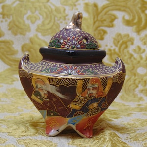 Könnte beinhalten: Ein kleiner, verzierter, dunkelvioletter und goldener Keramik-Räucherofen mit Deckel. Der Räucherofen ist mit komplizierten floralen und geometrischen Mustern verziert und zeigt zwei Figuren in traditioneller japanischer Kleidung.