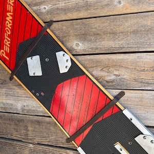 May include: A vintage Burton Performer snowboard featuring a red and black striped deck with the word PERFORMER in yellow text. The board includes black rubber traction pads, metal mounting plates, and two brown leather straps secured with metal rivets.