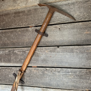 May include: A vintage wooden-handled ice axe with a metal head and leather straps. The axe is mounted on a wooden wall.