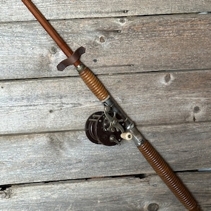 May include: A vintage wooden fishing rod with a brown leather handle and a metal reel. The rod is lying on a wooden surface.