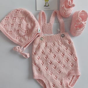 Könnte beinhalten: Ein Baby-Kleidungsset in zartem Rosa, bestehend aus einem Strampler mit Trägern, einer Mütze und zwei Paar Babyschuhen. Das Set ist mit einem zarten Lochmuster gestrickt. Das Etikett lautet "Esmeralda & Lana".