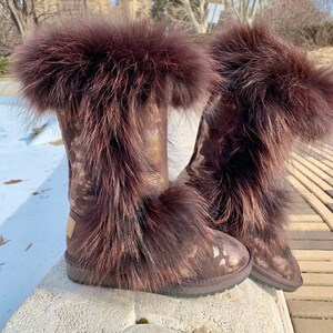 Puede incluir: Un par de botas de piel sintética marrón con un acabado metálico dorado y un estampado de camuflaje. Las botas tienen un ribete de piel esponjosa alrededor de la parte superior.
