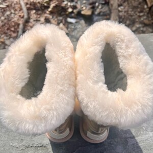 May include: A pair of beige winter boots with a soft, fluffy, cream-colored lining. The boots have a light pink sole and a metallic gold exterior. The boots are designed to keep feet warm in cold weather.